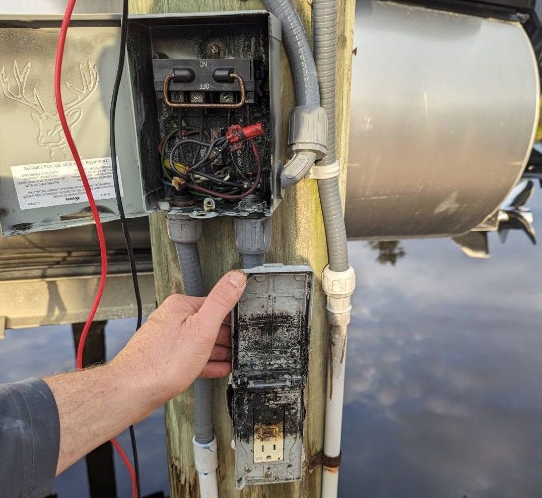 Hand pointing at an open, damaged electrical control box with exposed wiring on industrial equipment.