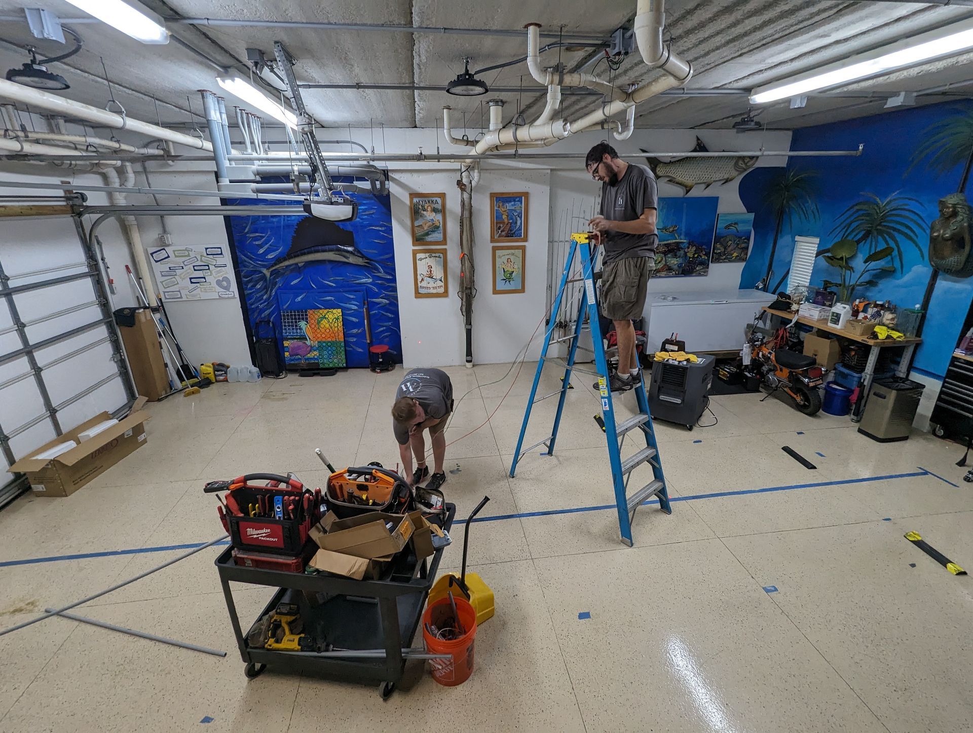 People painting colorful murals in a garage workshop, with ladders and supplies scattered around.