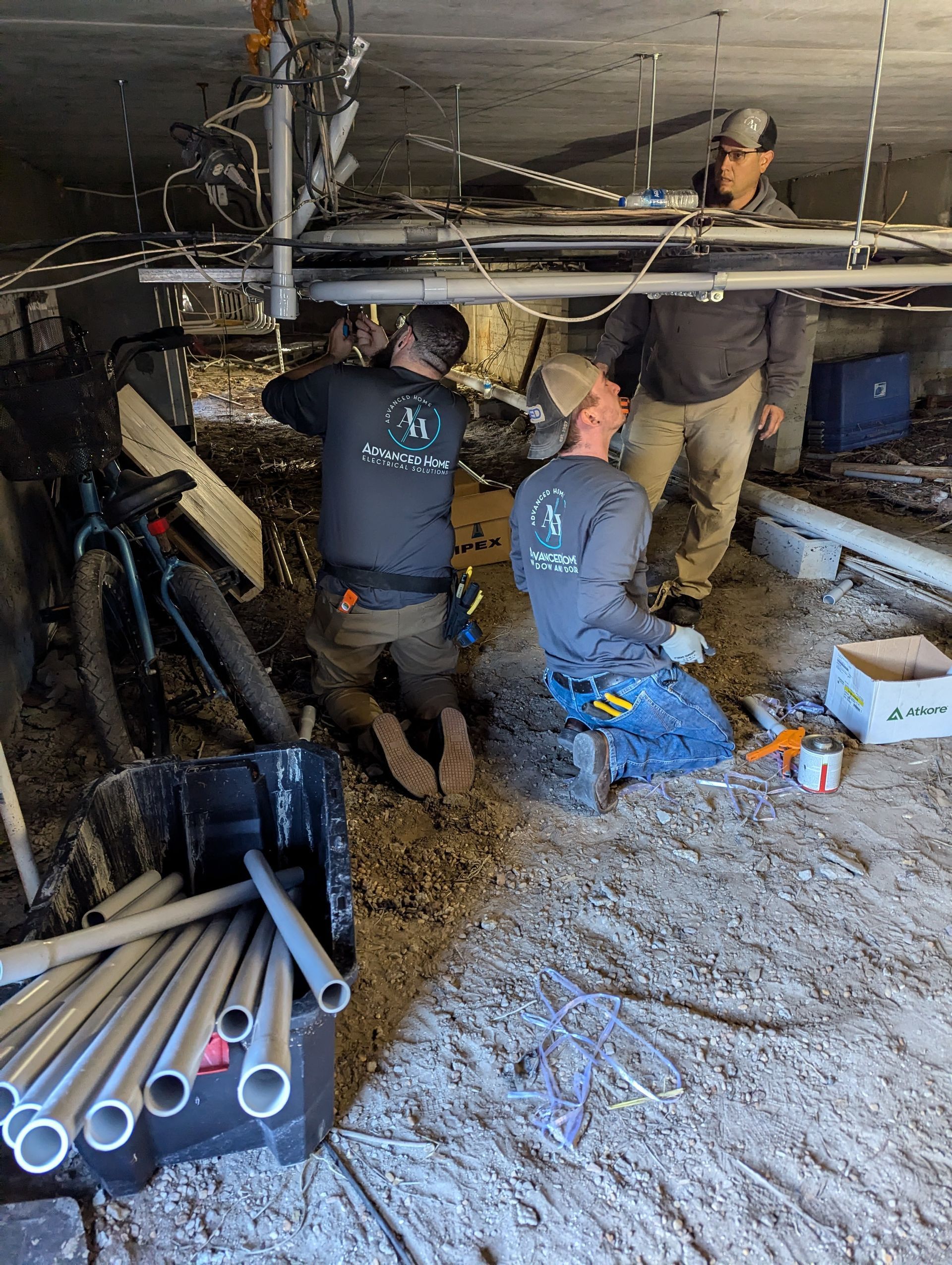 Workers repairing pipes in a muddy crawlspace beneath a building