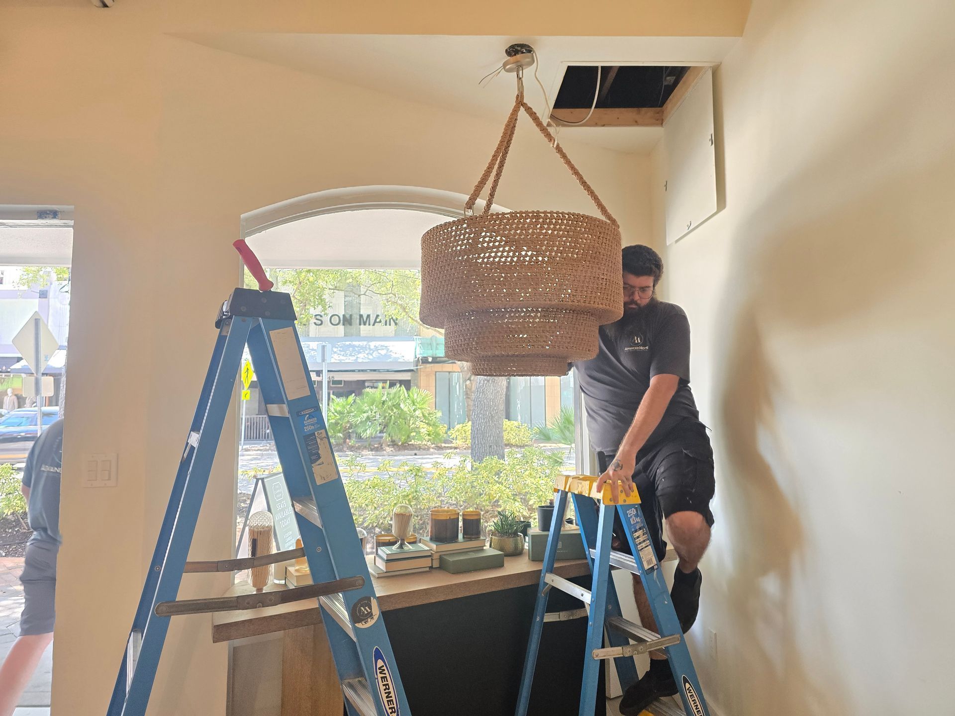 Two workers on ladders installing a large woven pendant lamp in a bright room