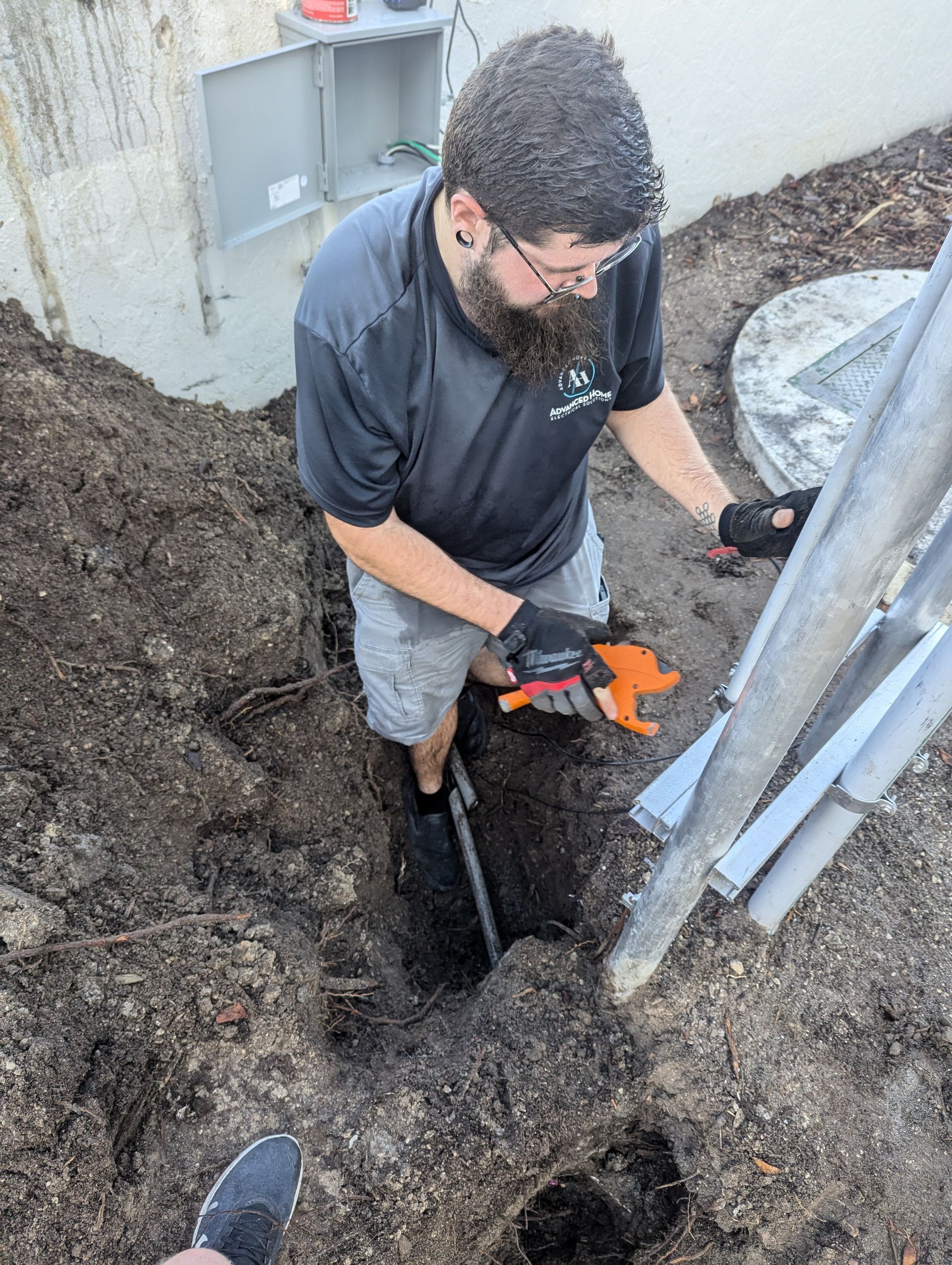 Worker in a trench using orange power tool beside utility pipes and exposed soil.