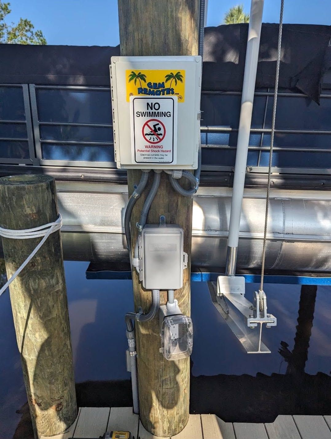 Warning sign and electrical box mounted on dock posts beside dark water and a metal barrier.