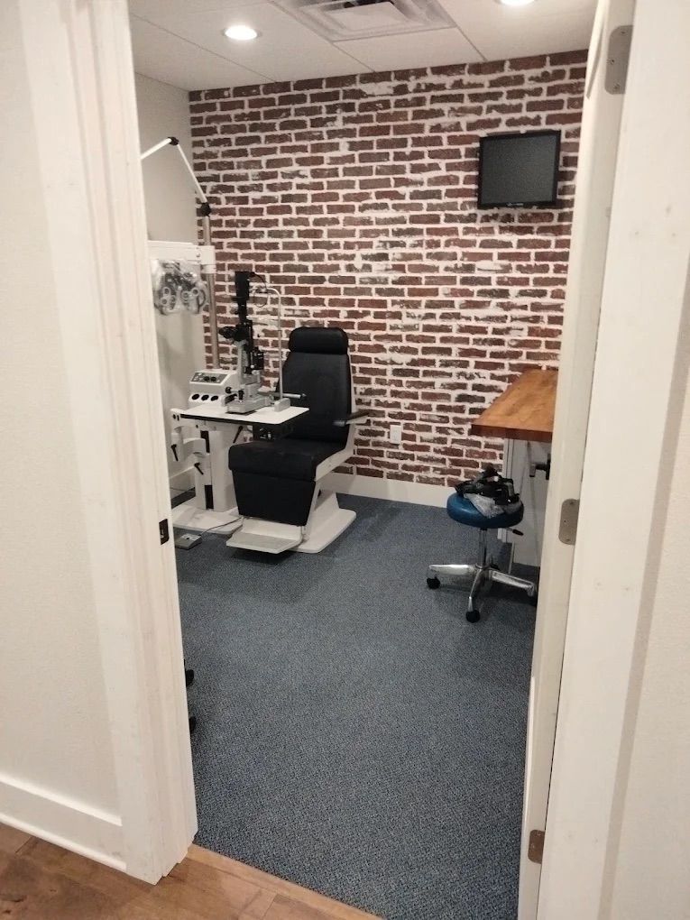 Small medical exam room with a black exam chair, brick accent wall, and overhead monitor.