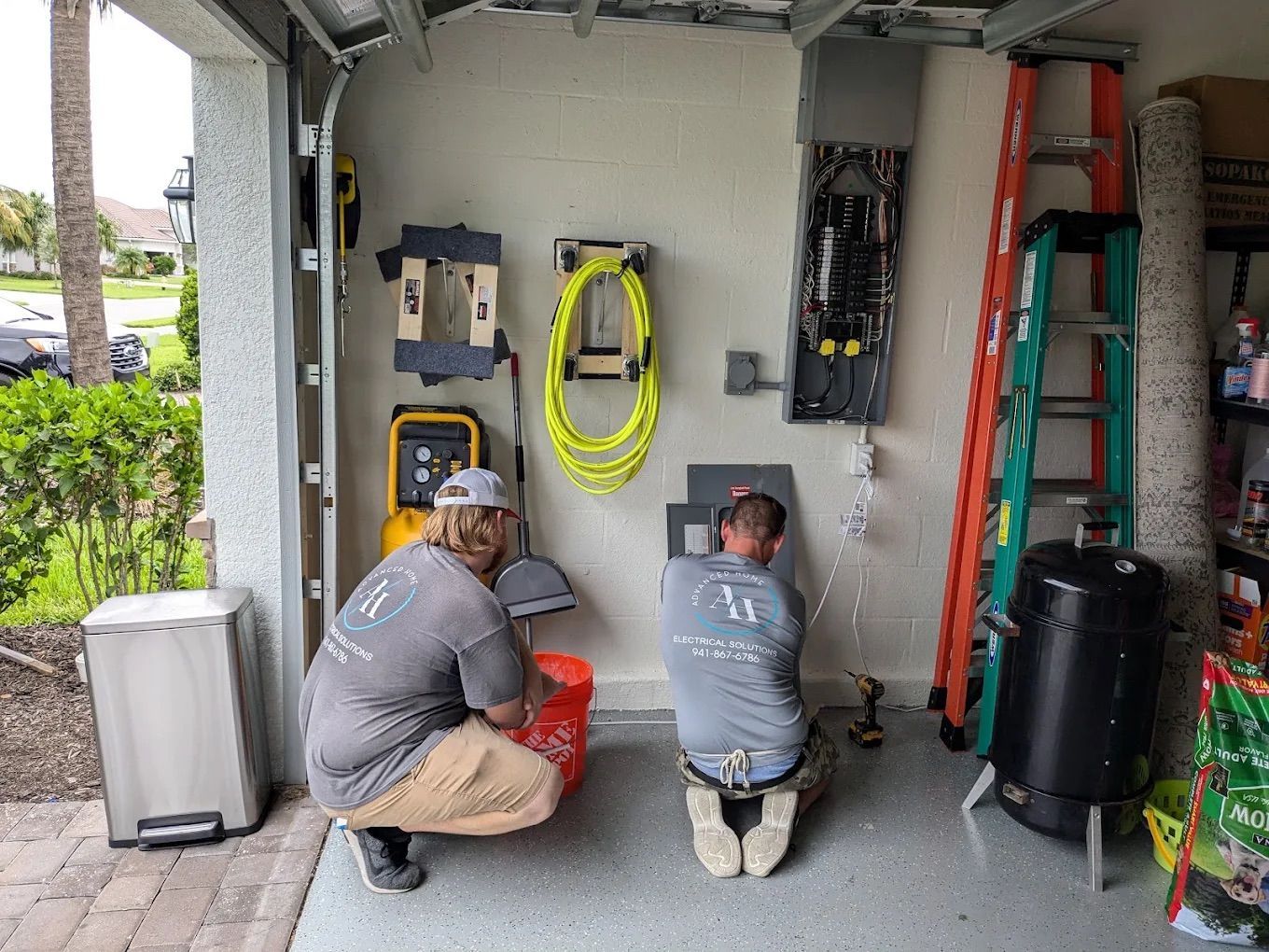 Two workers kneel in a garage, installing or repairing electrical wiring on a wall.