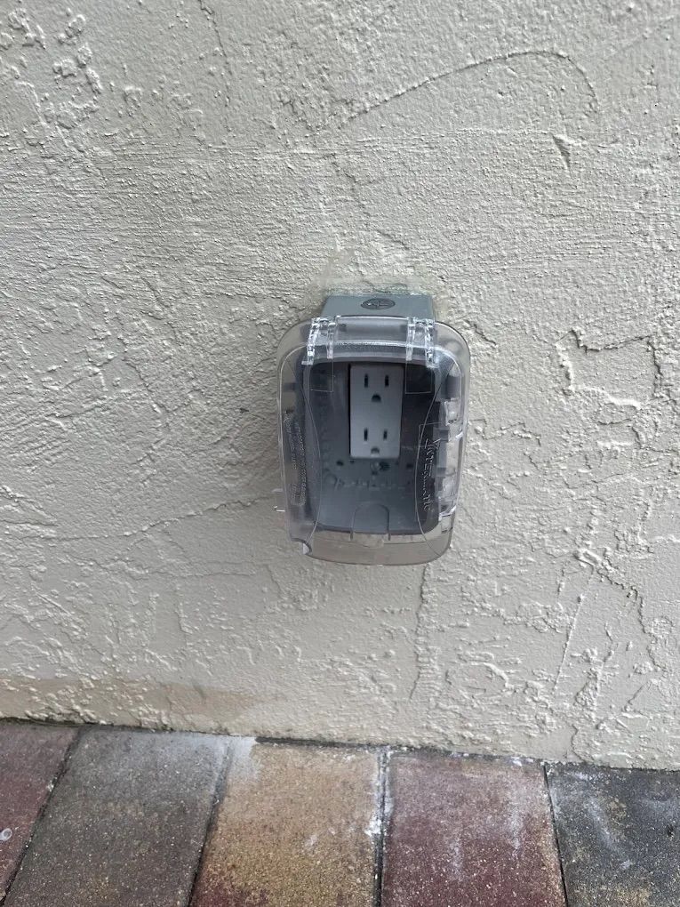 Outdoor electrical outlet in a beige stucco wall, covered in clear plastic.