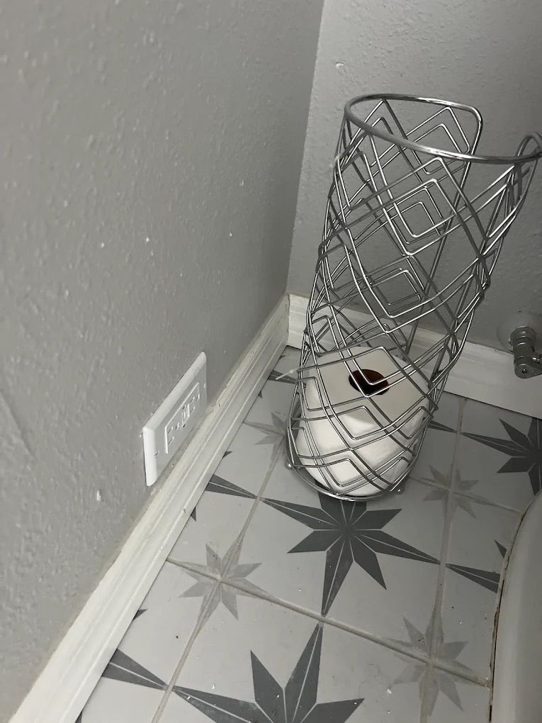 White wire trash bin holding a toilet paper roll, beside a wall outlet on a patterned tile floor.