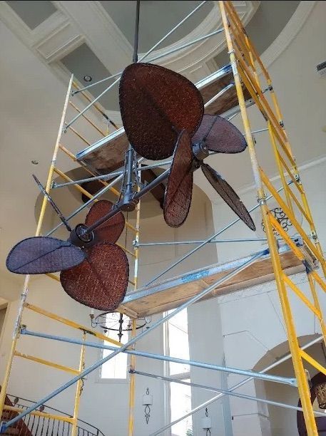 Large hanging sculptural fan installation with blue metal framework and brown mesh blades in an indoor atrium