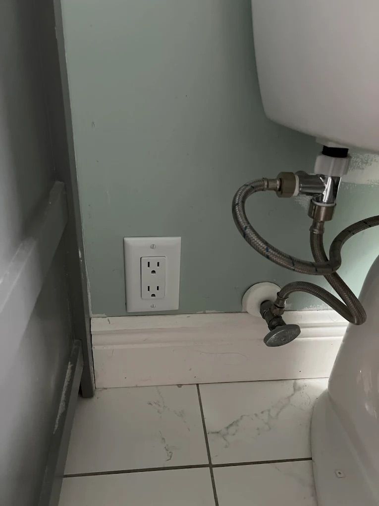 Wall outlet beside sink plumbing in a small bathroom with cracked tile floor and gray wall.