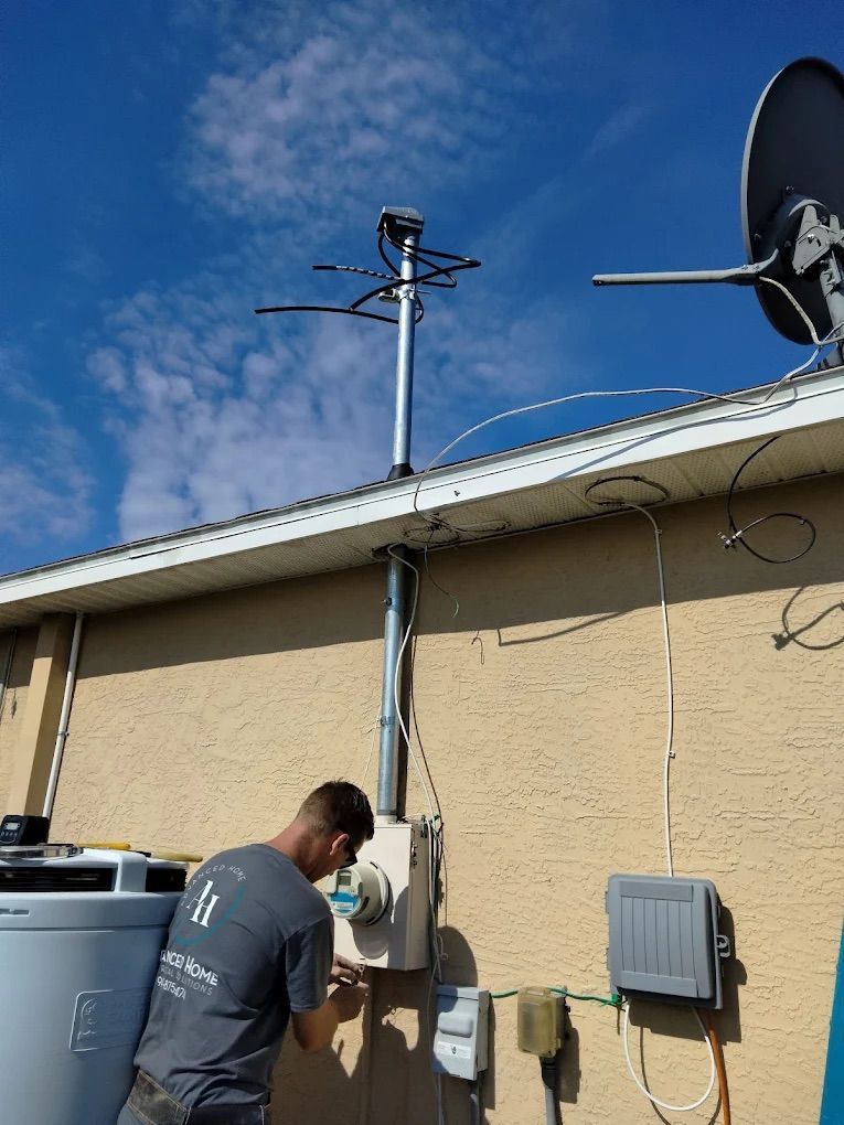Technician working on wall-mounted electrical equipment beside a house, with a roof antenna and satellite dish above.