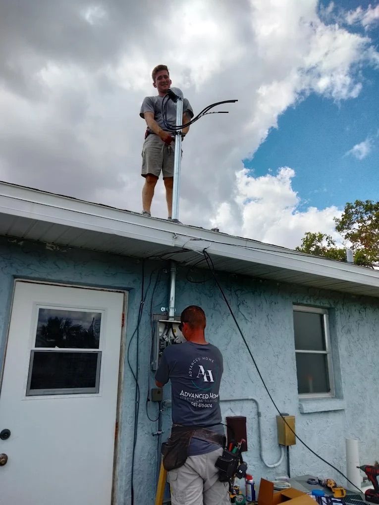 Two workers repair cables on a house roof and wall under a cloudy sky.
