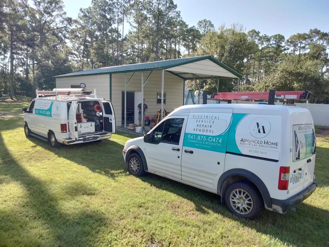 Two white service vans parked outside a small white building in a grassy, wooded area.