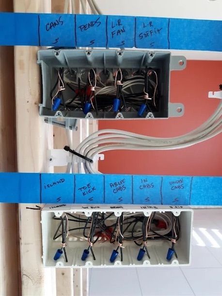 Open electrical boxes with labeled blue tape and exposed wires in a wall panel.