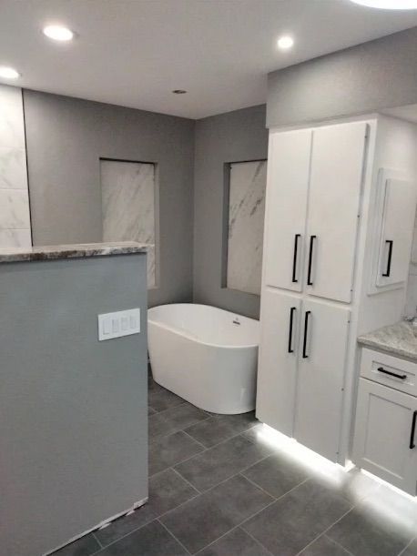 Modern bathroom with gray tile, white cabinetry, and a freestanding tub in a corner.