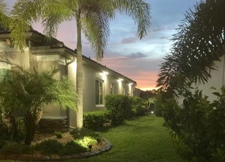 Tropical garden at dusk beside a white house, with palm trees and a pink-blue sunset sky.