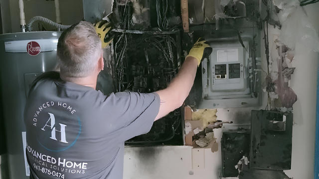 Worker inspecting electrical panel in a smoke-damaged room, wearing gloves and pointing at wiring.