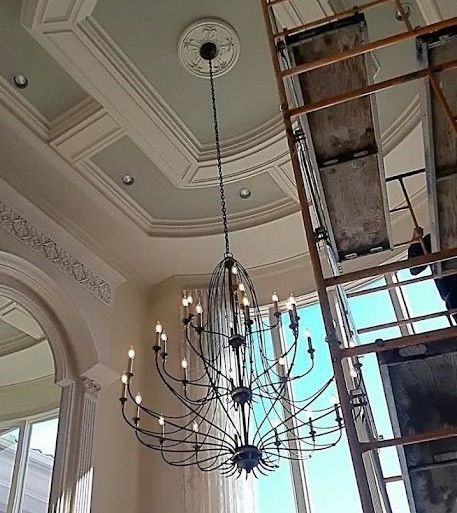Chandelier hanging from a ornate ceiling beside a tall scaffolding or ladder in a bright interior room
