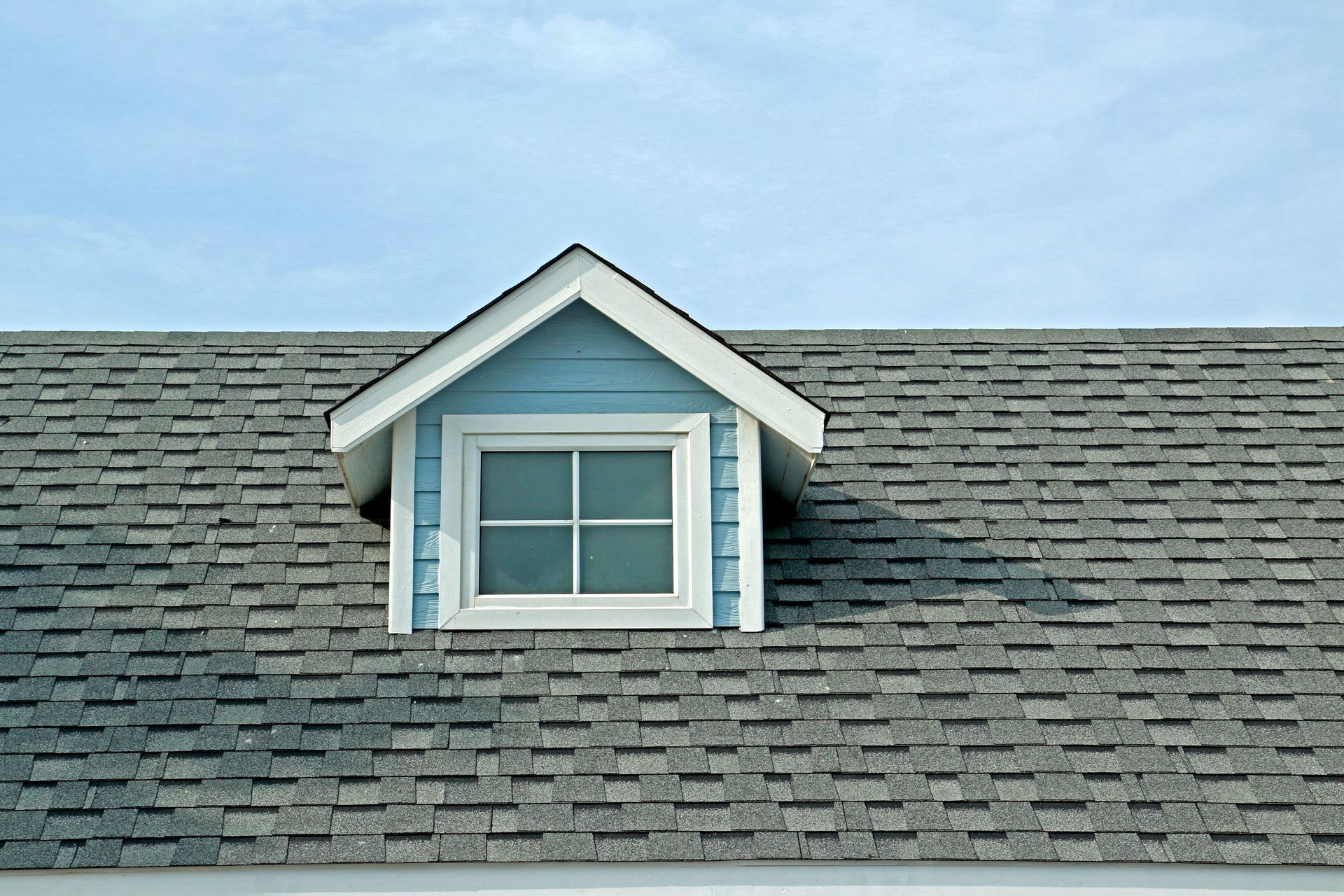 Blue dormer with white trim and window on a gray shingled roof against a blue sky.