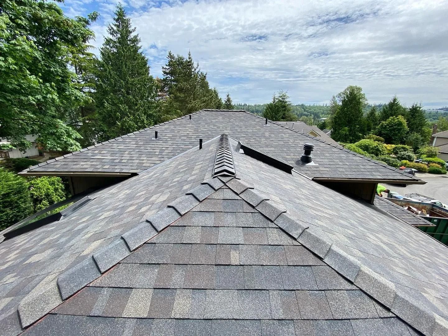 View of a roof with gray shingles and ridge cap, surrounded by trees under a cloudy sky.
