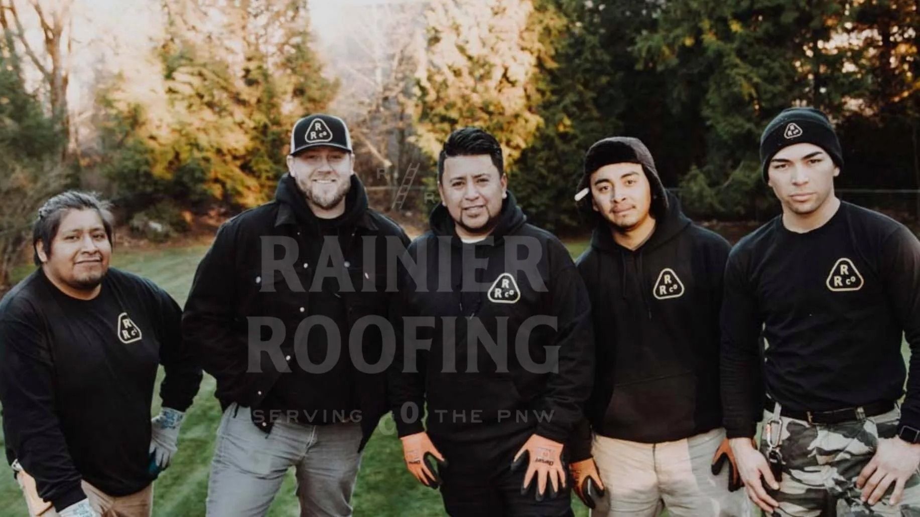 Rainier Roofing crew outdoors. Five men in black shirts with logo, standing, smiling, with a green backdrop.