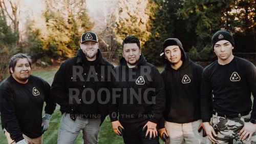 Rainier Roofing crew outdoors; five men in black shirts with logo, posing in front of foliage.