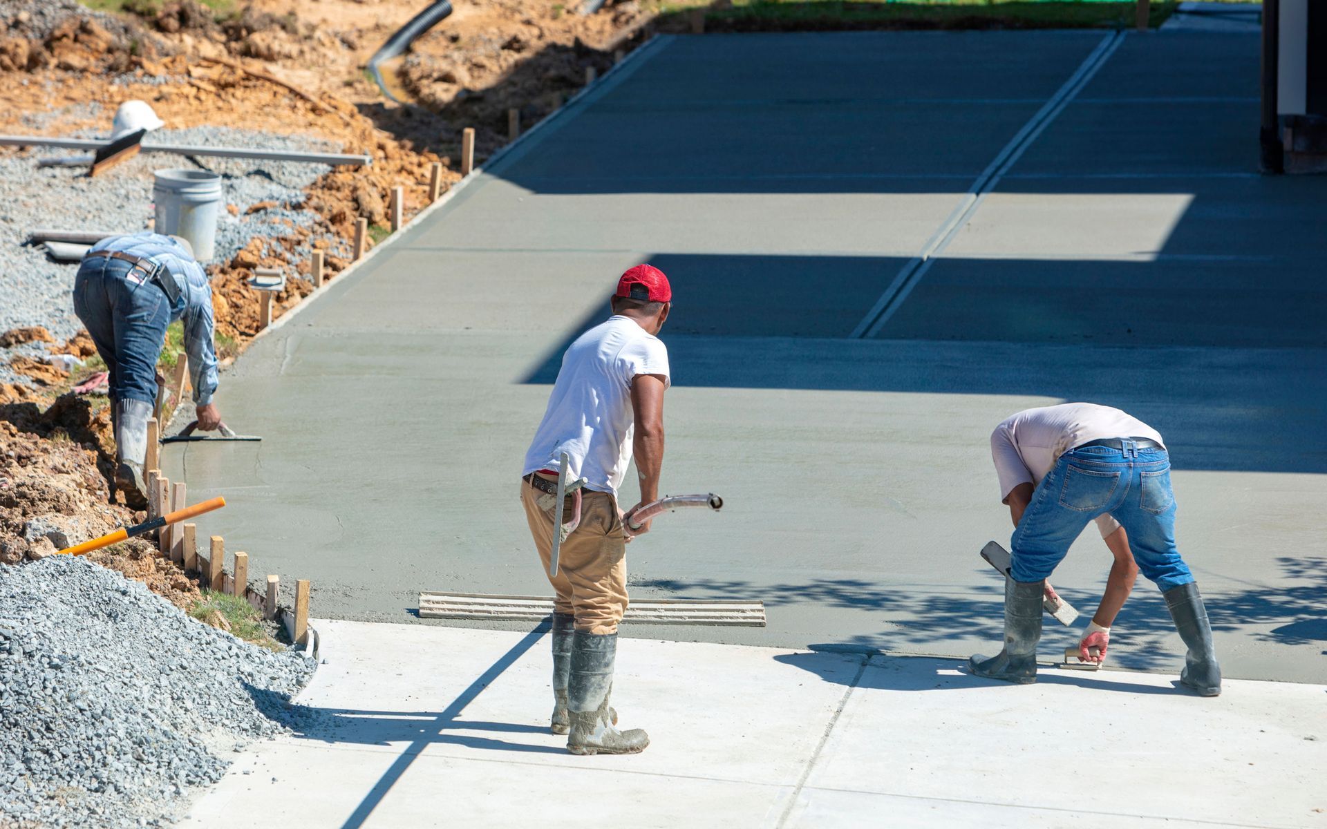 Three construction workers smoothing a newly poured concrete driveway with tools.