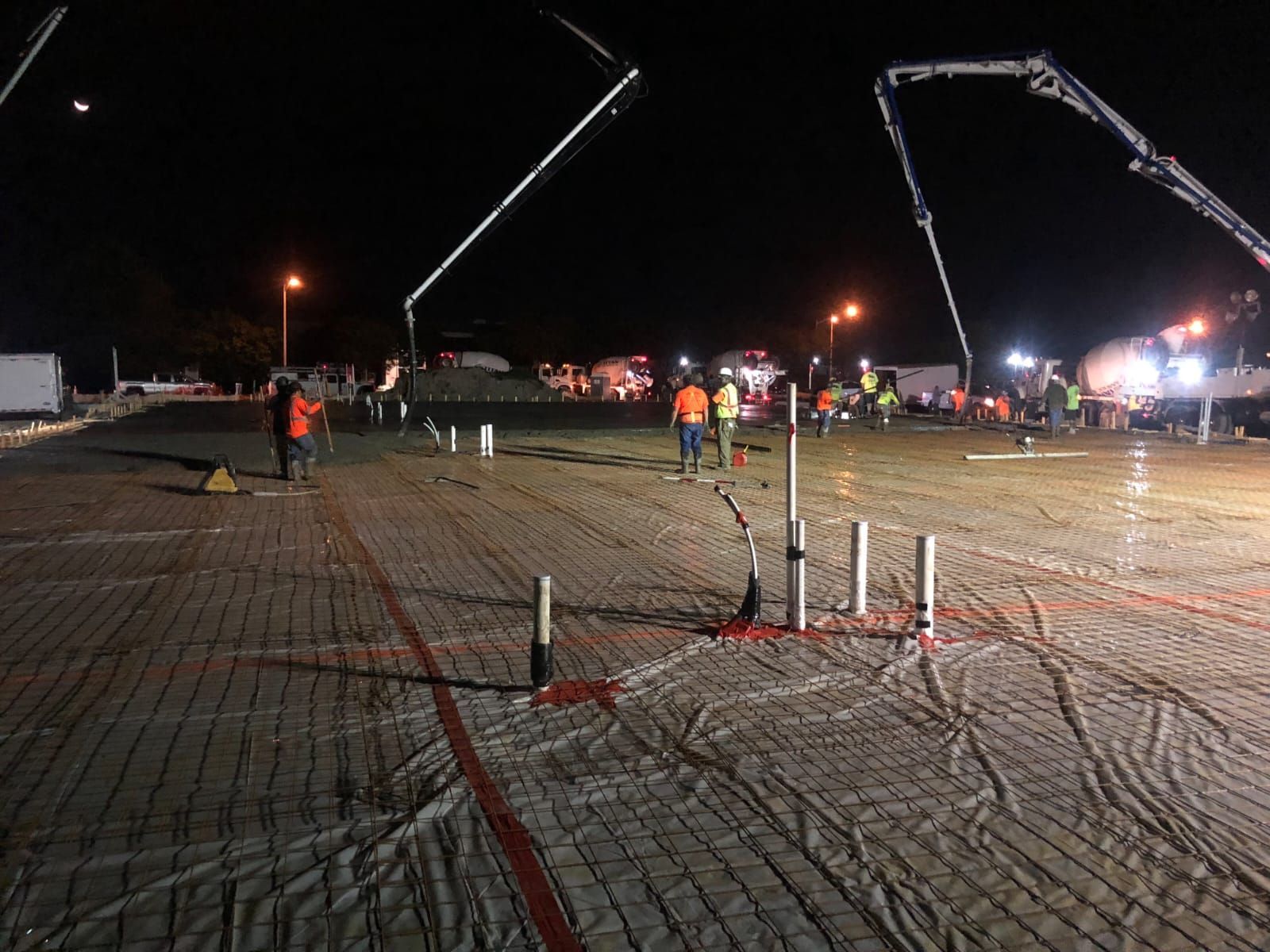Nighttime construction site with concrete being poured. Workers, pumps, and lights are visible.