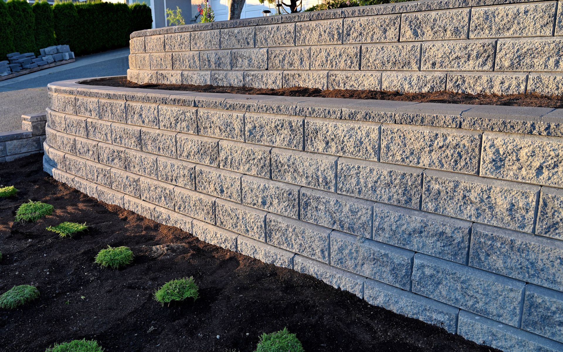 A retaining wall constructed of gray textured blocks, holding soil and small green plants.