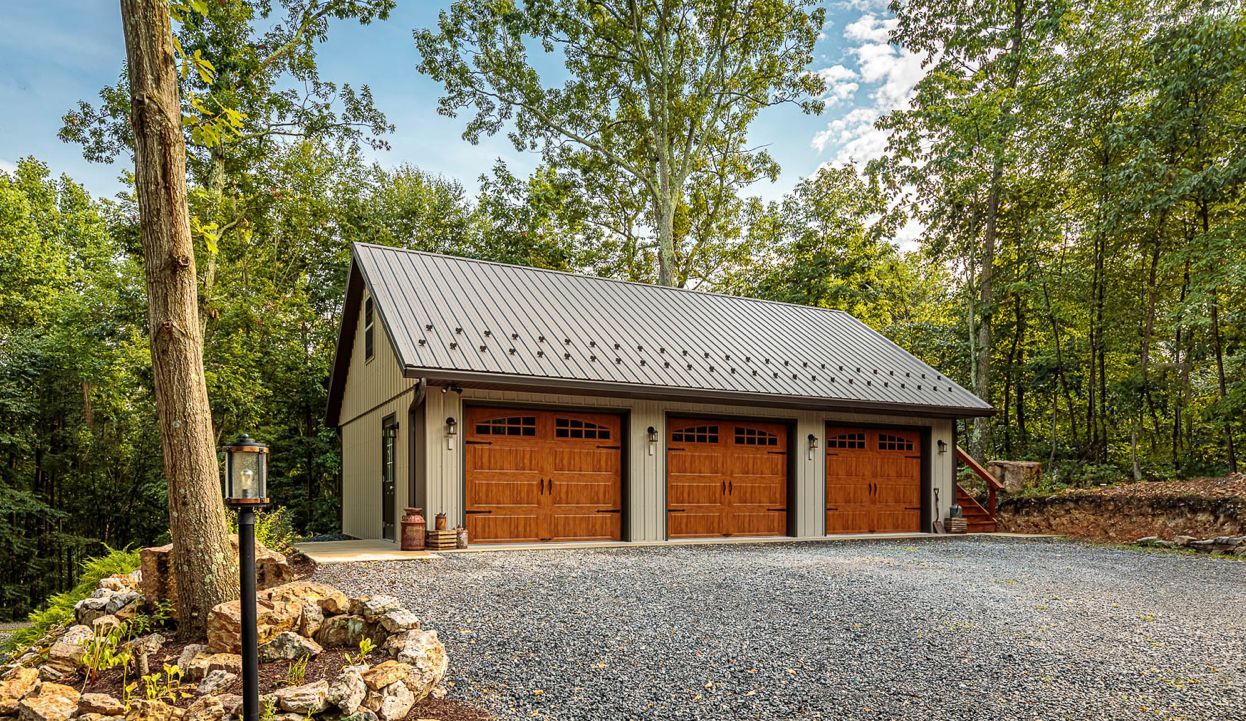 Two-car garage with wooden doors and a gray shingled roof in a wooded gravel driveway