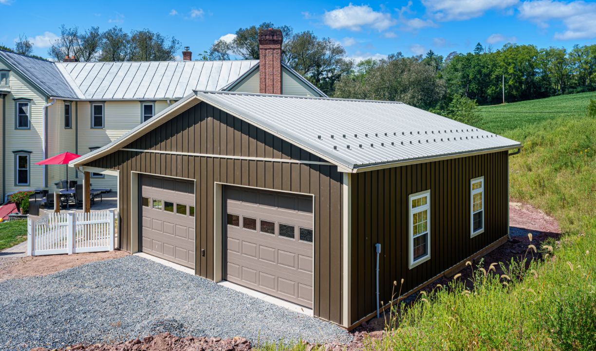 Brown two-car garage with white metal roof beside a house on a grassy rural hillside.