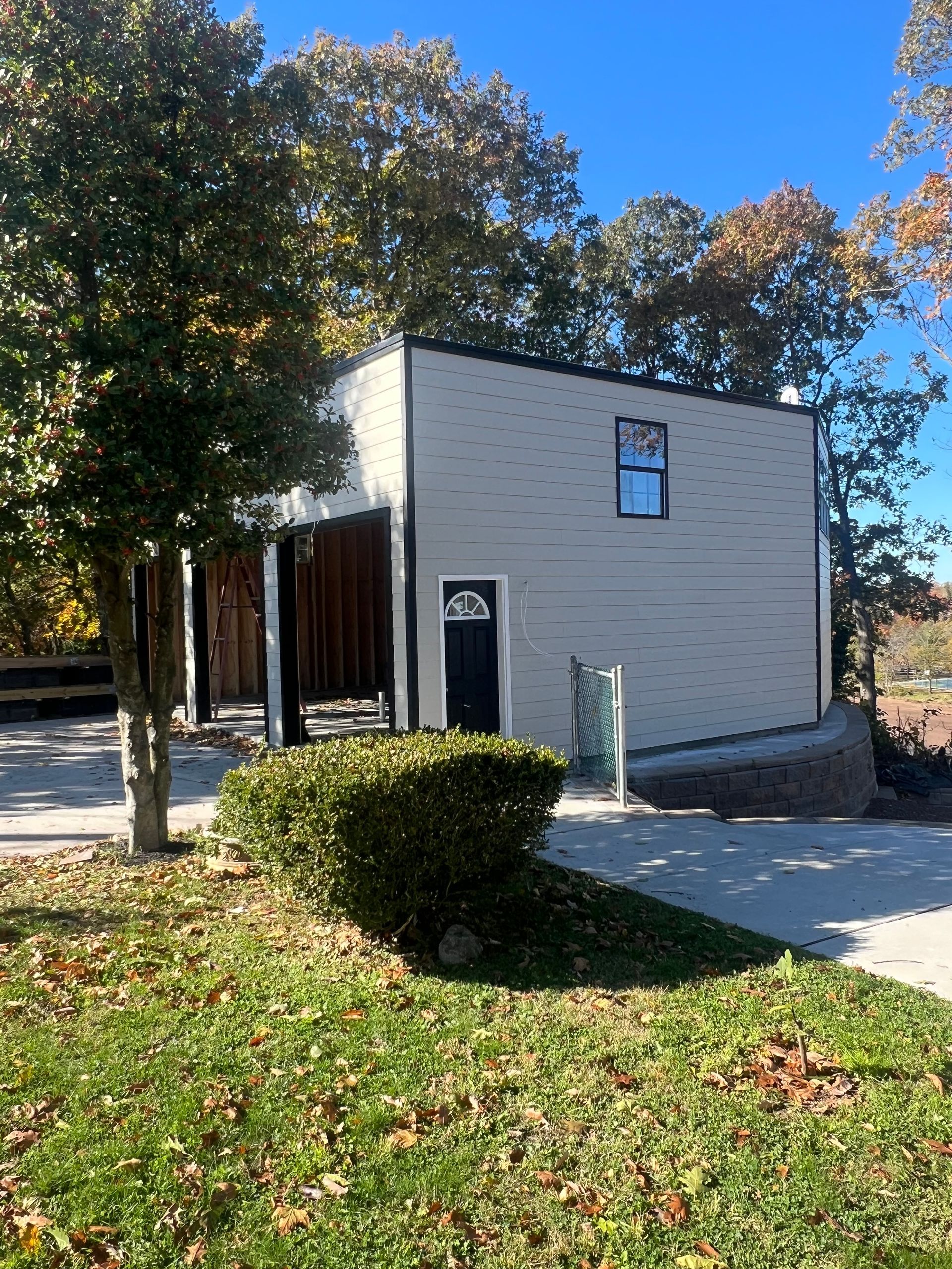 Small white building with dark trim, a door and window, surrounded by trees and grass in autumn sunlight