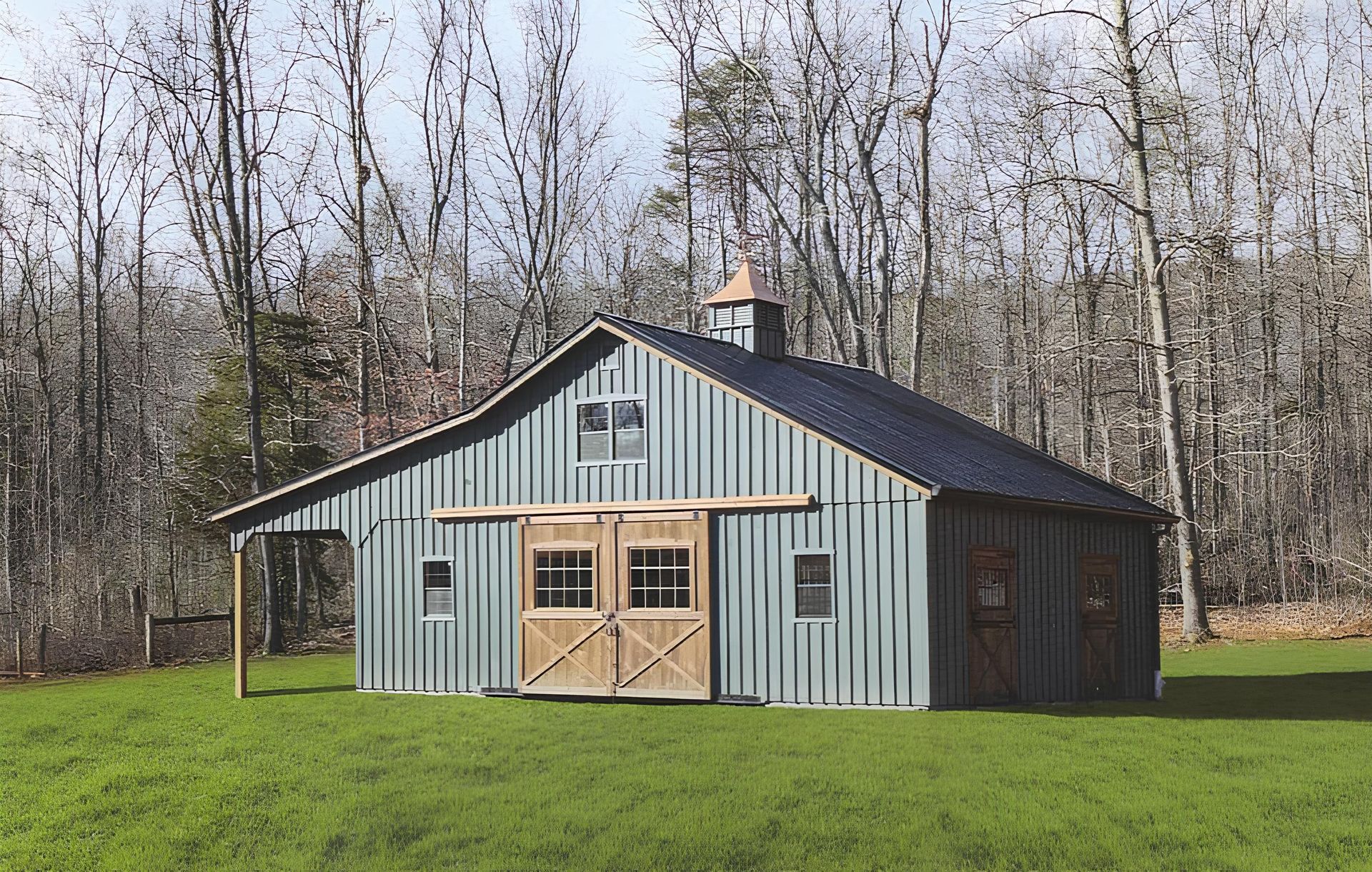 Small gray metal barn with tan double doors on a grassy field, backed by leafless trees.