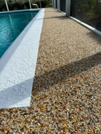 Pebble walkway beside a turquoise swimming pool, bordered by glass walls and a shadowed path.