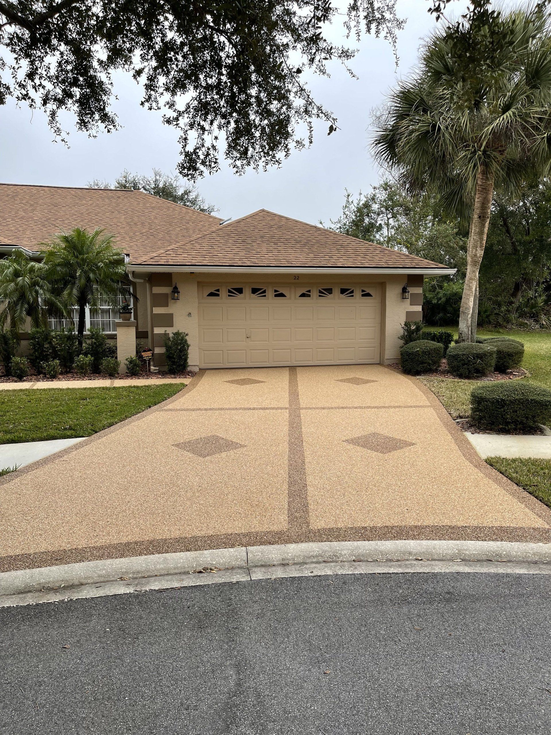 Suburban house with a tan garage, stone driveway, and trimmed hedges under trees