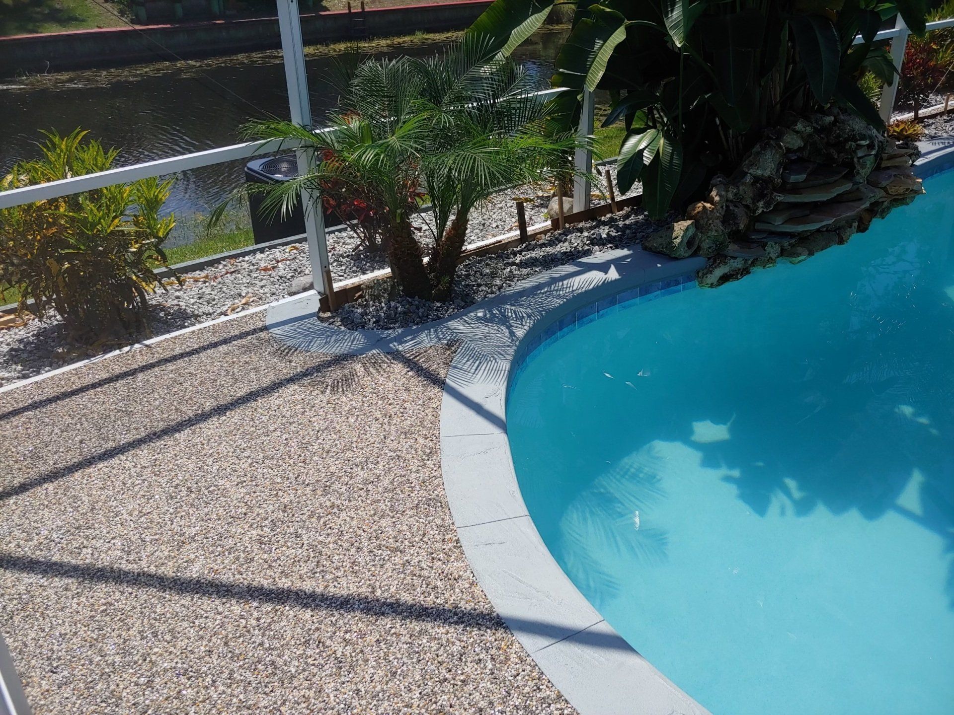 Curved swimming pool edge beside gravel patio, white railing, and tropical plants in bright sunlight