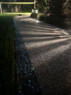 Sunlit gravel path curving through a garden, bordered by bushes and grass with dappled shadows.