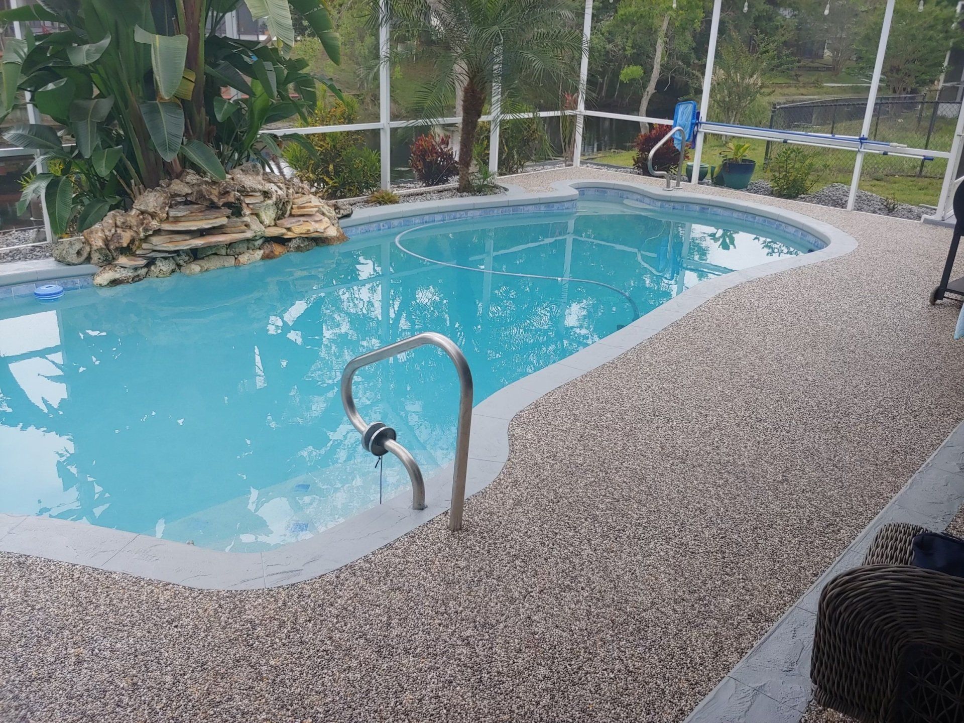 Backyard pool with turquoise water, pebble deck, tropical plants, and a person cleaning near the far end
