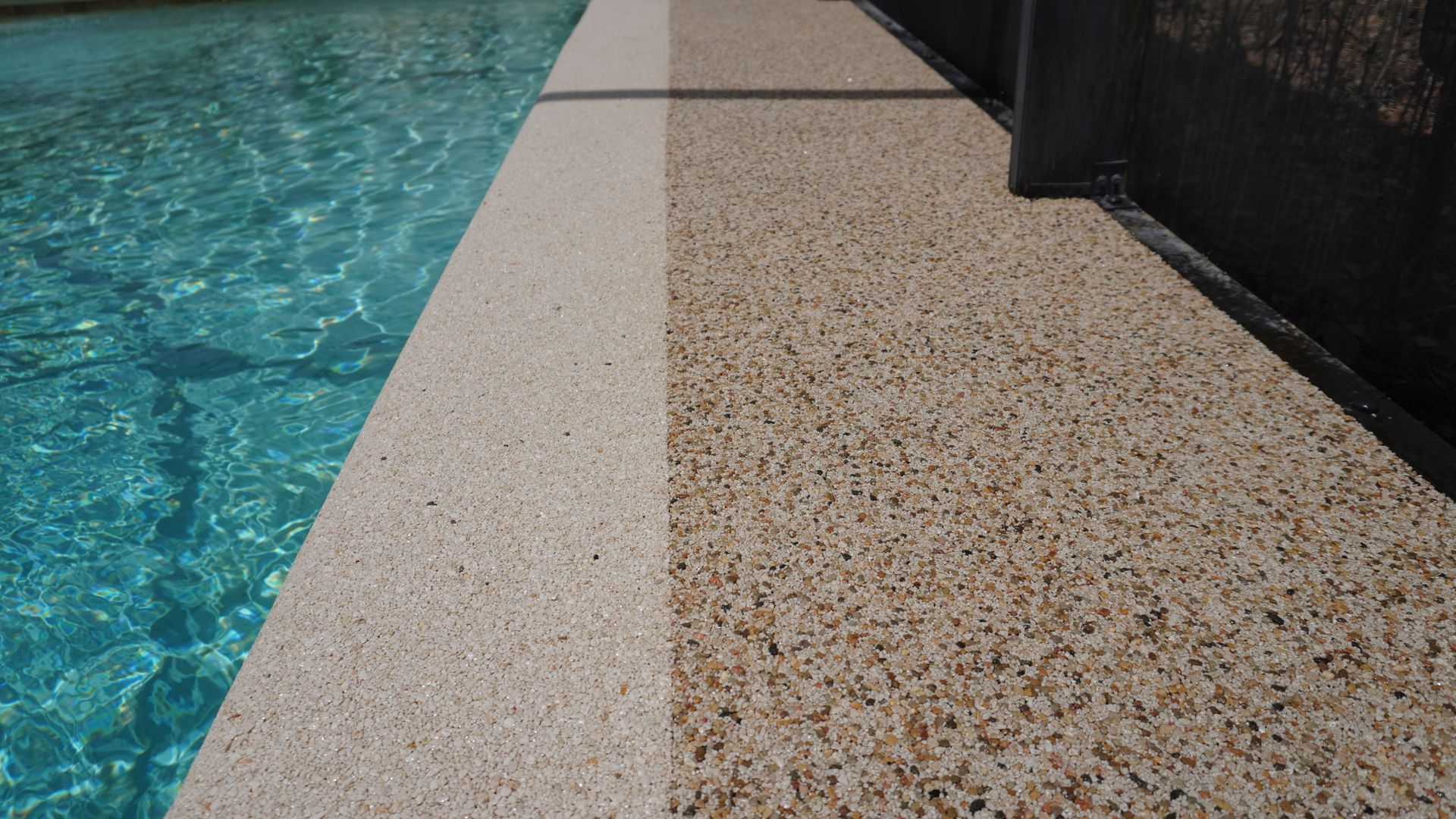 Swimming pool edge with blue water and a beige textured deck beside a dark fence