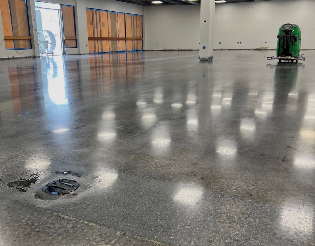 Shiny empty warehouse floor with reflections and a floor drain; green floor machine at right.