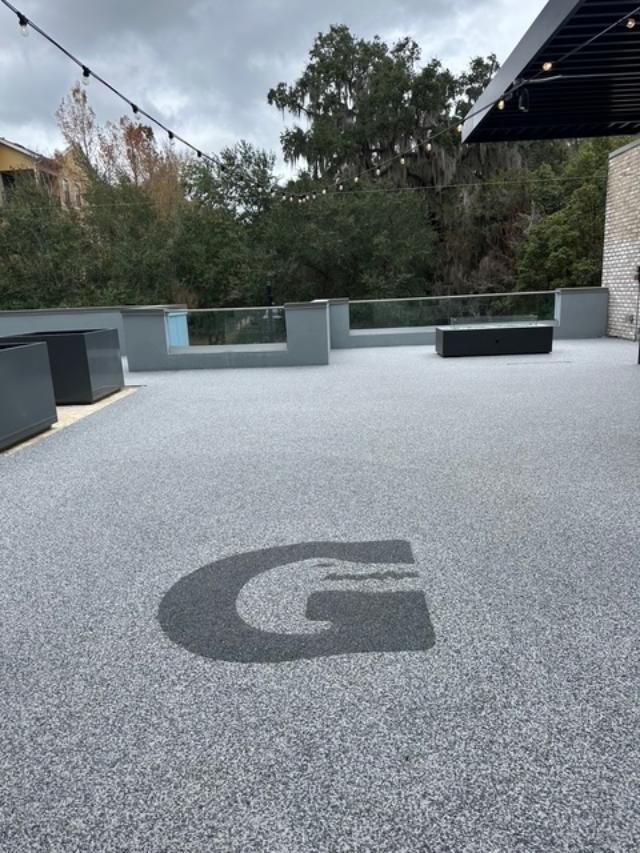 Outdoor terrace with gray concrete flooring and a large black “G” logo, bordered by low seating and trees.