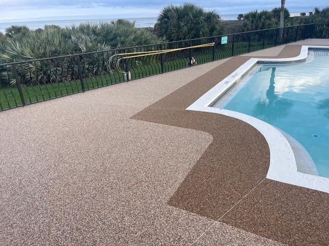 Curved pool deck with textured tan and brown surface beside clear blue water and a black fence.