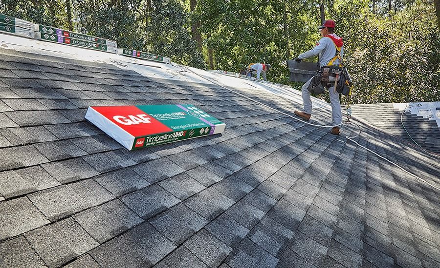A roofer on a rooftop with a bundle of shingles. The shingles are grey. Green trees are in the background.