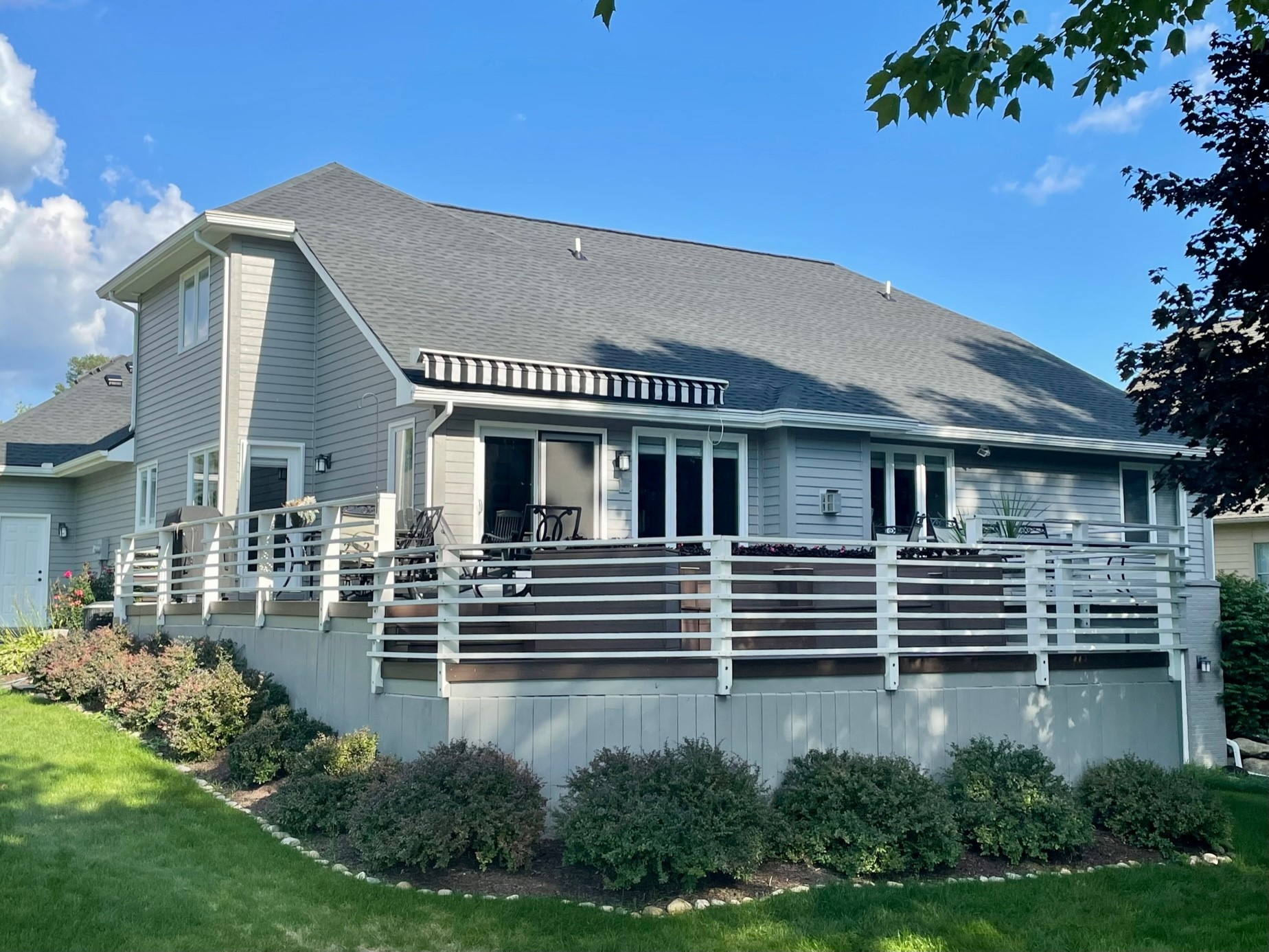 Grey house with large deck and white railing, lush landscaping and blue sky.