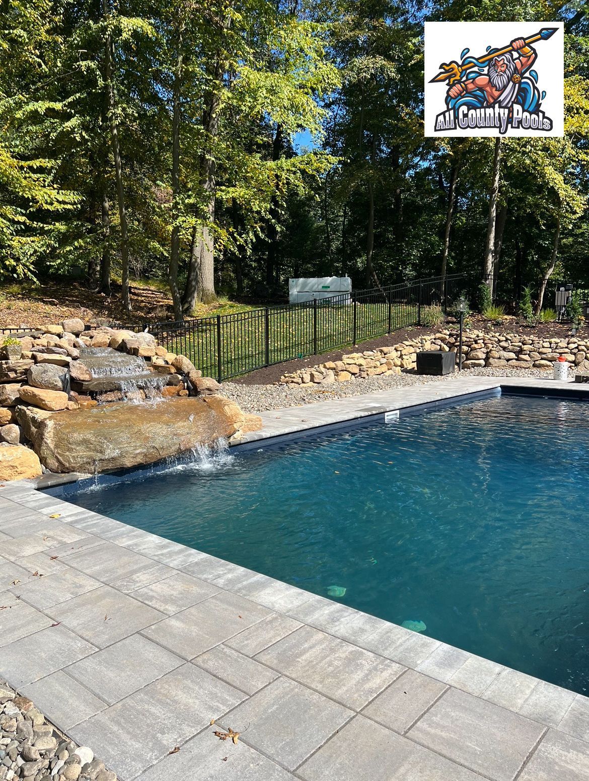 A large swimming pool with a waterfall in the backyard surrounded by trees.