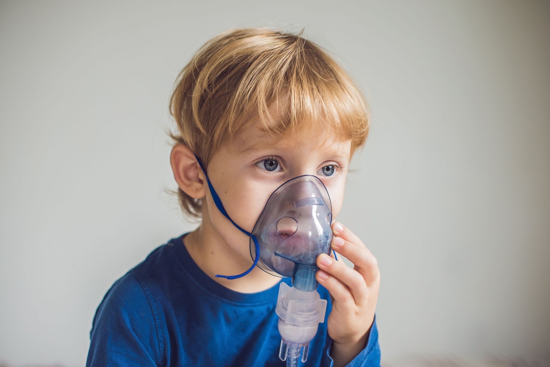 Child using a nebulizer mask, wearing a blue shirt against a plain background.