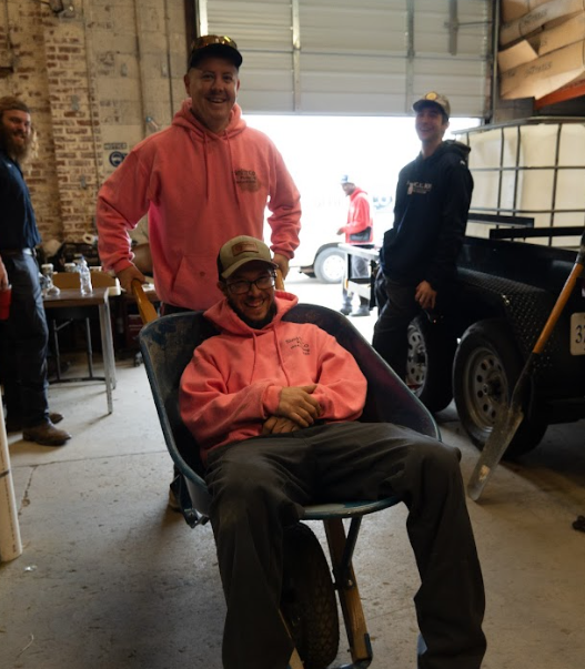 A man in a pink hoodie is sitting in a wheelbarrow