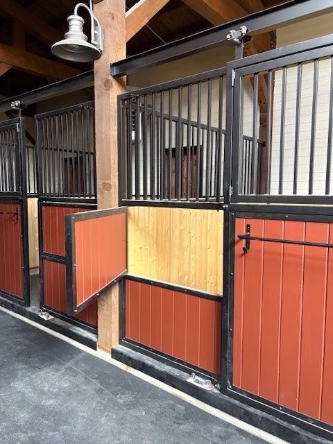 Horse stalls with red and yellow panels, black bars, and open doors under a wooden beam.