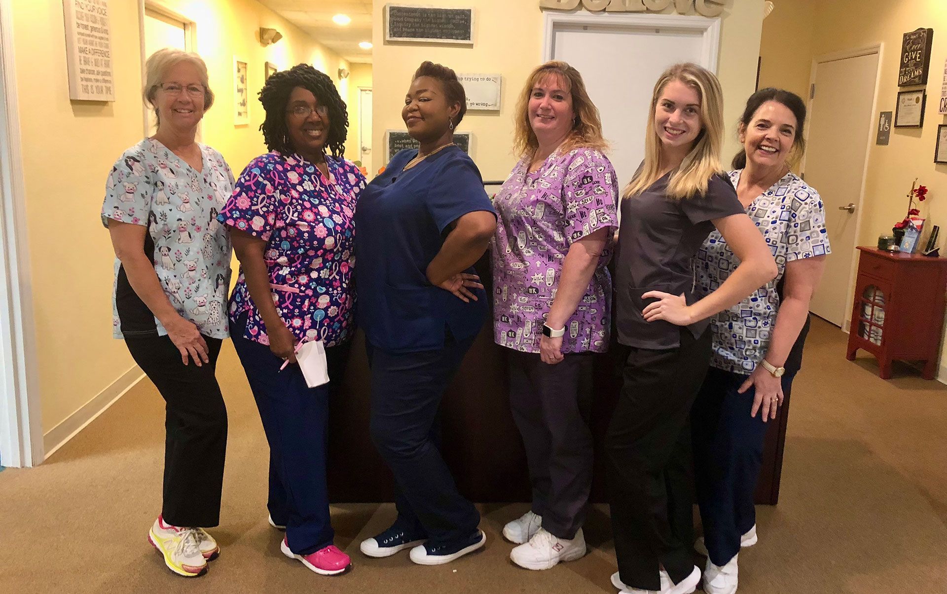 Six women in scrubs posing for a photo, standing in a room with beige walls and a desk.