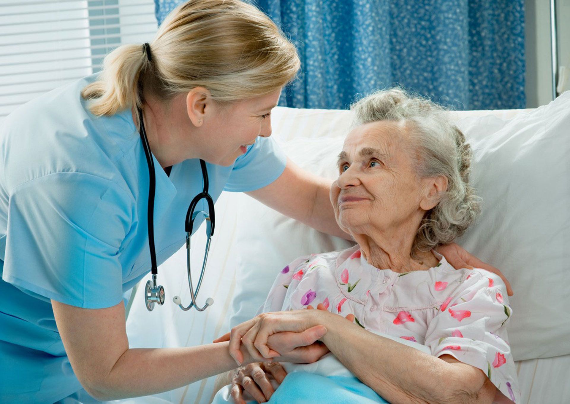 Nurse in blue scrubs smiles, holds hands with elderly woman in floral gown, in a hospital room.