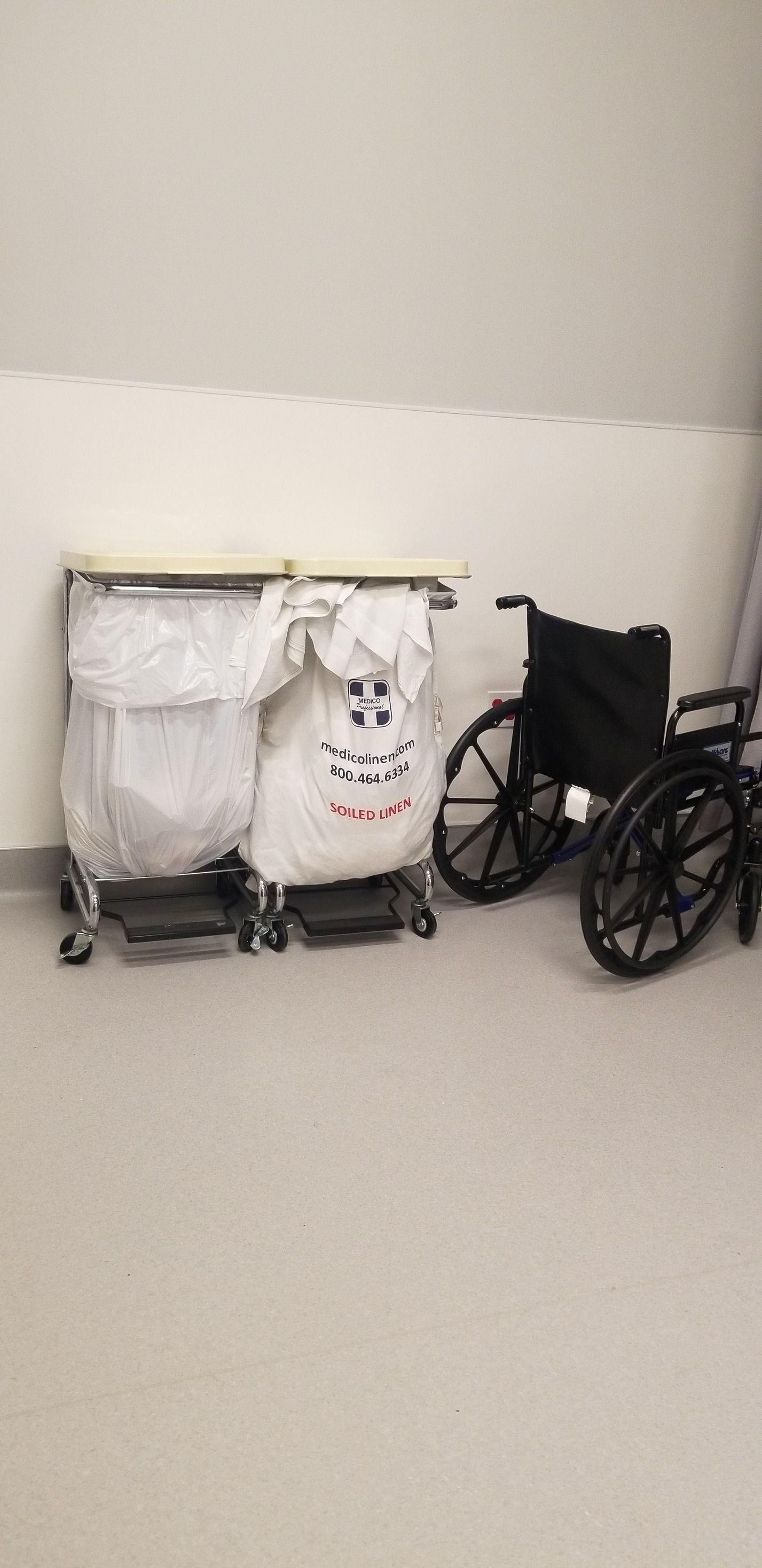 A wheelchair and two laundry carts in a sterile room. Carts have white bags. Gray and white walls and floor.