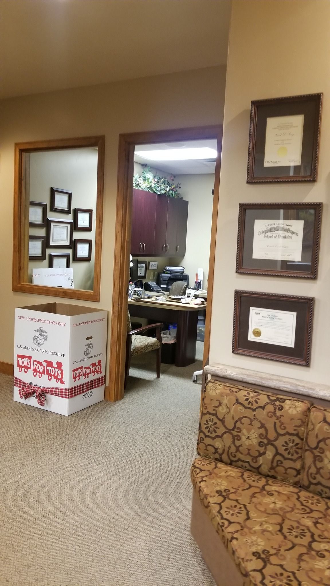 Hallway with office entrance, framed certificates, large box, mirror with documents, and patterned bench.